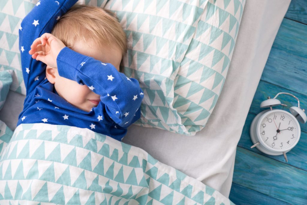 Picture of a sleeping toddler in bed, next to a white alarm clock.